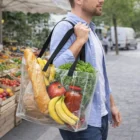 Man carrying a Crystal Clear Stadium Tote Bag 15L with bread, bananas, lettuce, and a tomato.