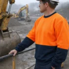 Man in Two-Tone Safety Windcheater Jacket and helmet stands at construction site near excavators.