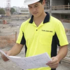 Man wearing Mens Fashion Truedry Safety Apparel Polo reviews blueprints at construction site.