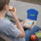 Boy eating a sandwich on a bench with his Sandwich Lunch Boxes, apple, and grapes nearby.