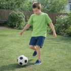 Boy in green Comfortfit Kids Regular Cotton Tee and denim shorts playing soccer on grass.