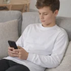 Teen boy in a personalized long sleeve T shirt sits on a couch, looking at his smartphone.