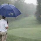 Golfer with a 76Cm Quality Promotional Golf Umbrella stands on a rainy course near a yellow flag.