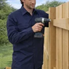 Man in a Drill Shirt Long Sleeve uses a power drill to fix a wooden fence outdoors.