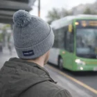 Person in Acrylic Beanies With Pompon waits at bus stop as a green bus arrives.