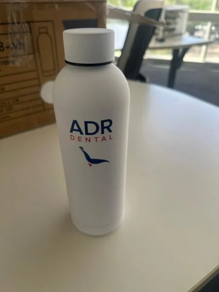 White ADR Dental water bottle—perfect for gifts for events—on a round white table, boxes behind.