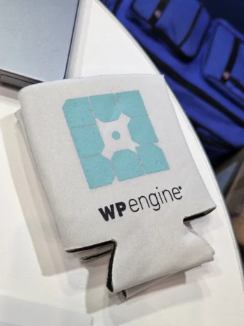 A white drink koozie with the WP Engine logo, printed from an EPS logo file format, on a table.