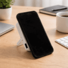 A smartphone on a stand sits on a wooden desk with a keyboard, laptop, and coffee.