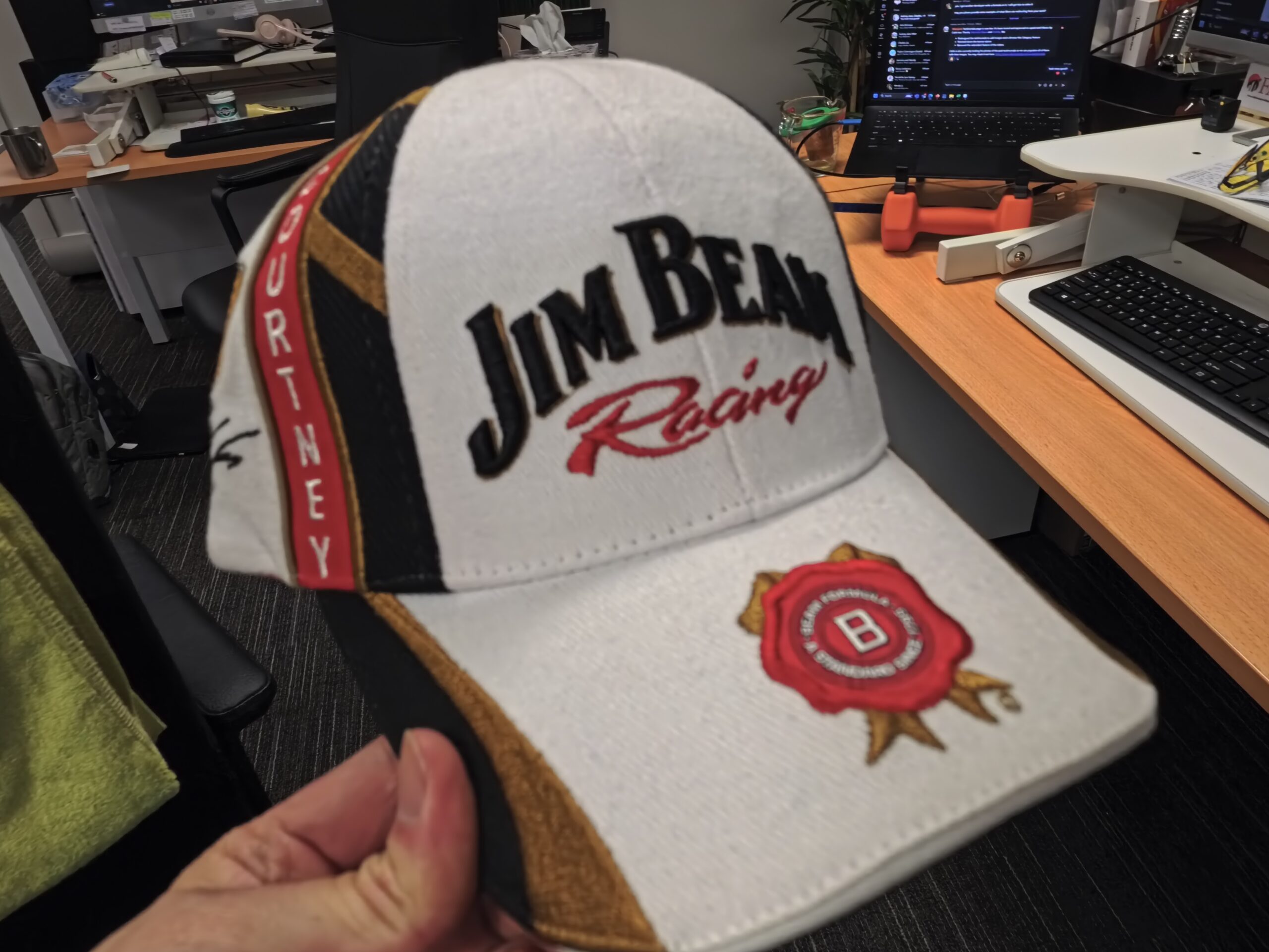 A hand holds a white Jim Beam Racing cap, showcasing brand consistency in an office setting.