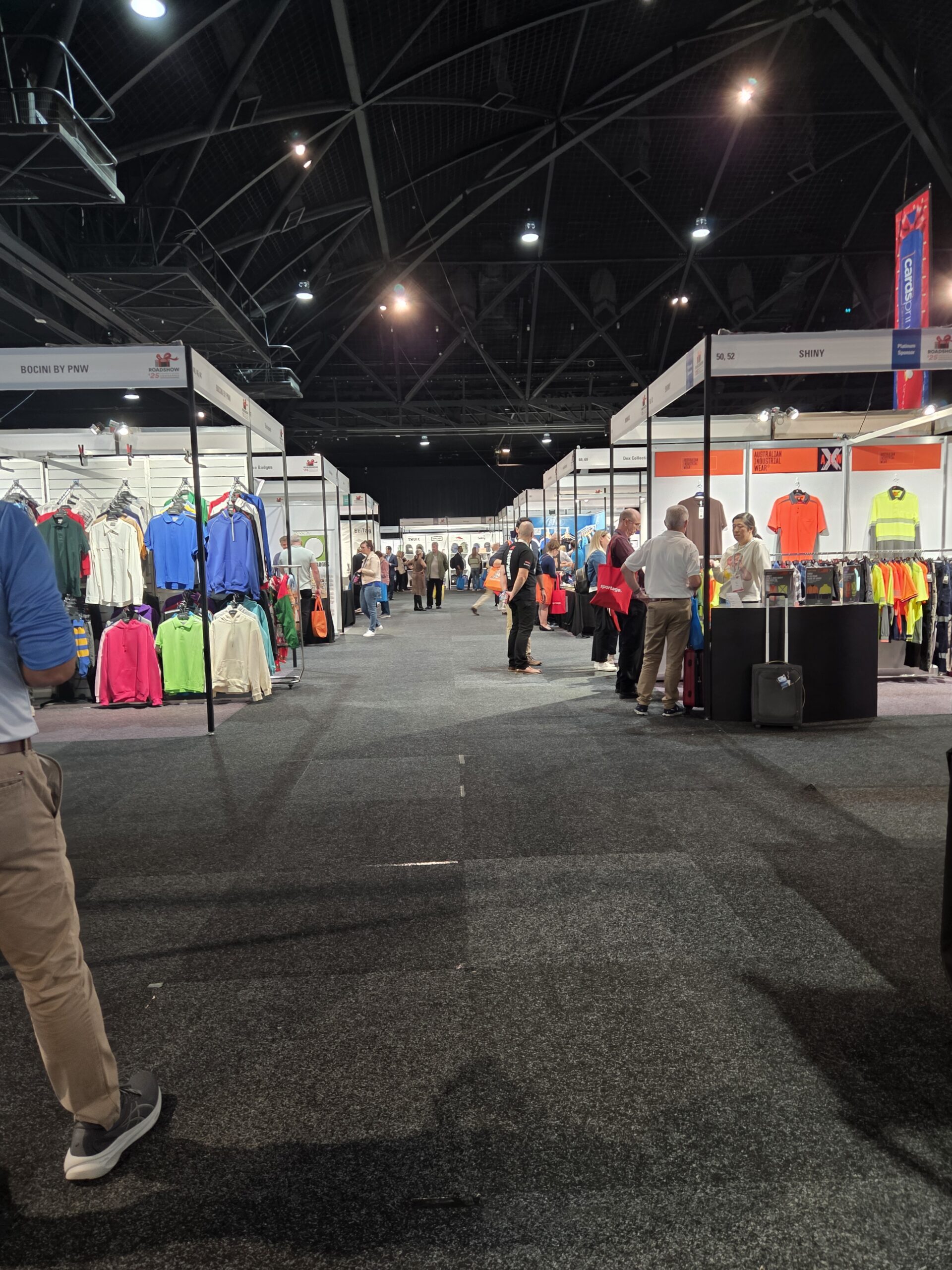 People walk an indoor expo for multi-day events, browsing clothing booths under bright lights.