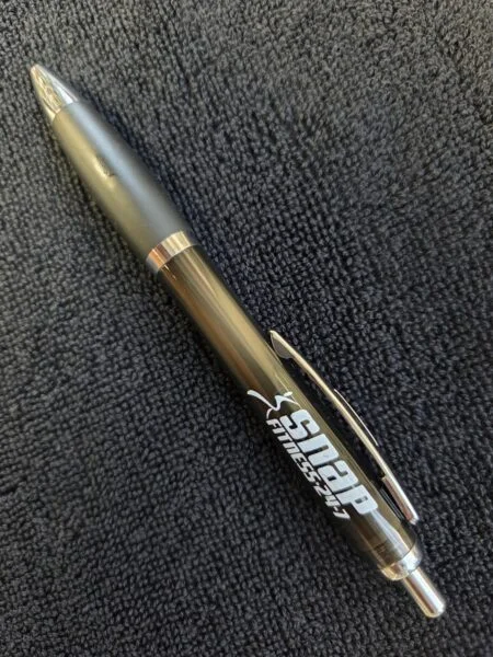 Black SNAP Fitness pen, perfect for heavy use, on a dark textured fabric background.