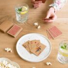 Person playing cards at a wooden table with drinks, popcorn, and scattered playing cards.