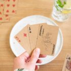 Hand holding three playing cards: 3 of diamonds, ace of spades, and joker, over a table.