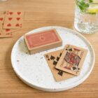Playing cards on a white plate, with a drink and more cards on a wooden table.