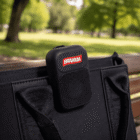 A portable black BRAHMA speaker clipped to a black bag on a park bench.