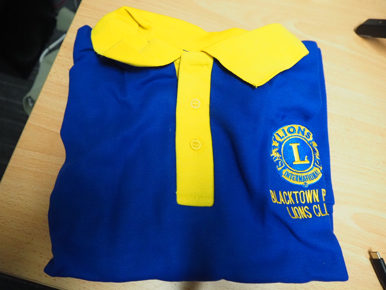 Folded blue and yellow Lions Club polo shirt, ideal for various print methods, on wood surface.