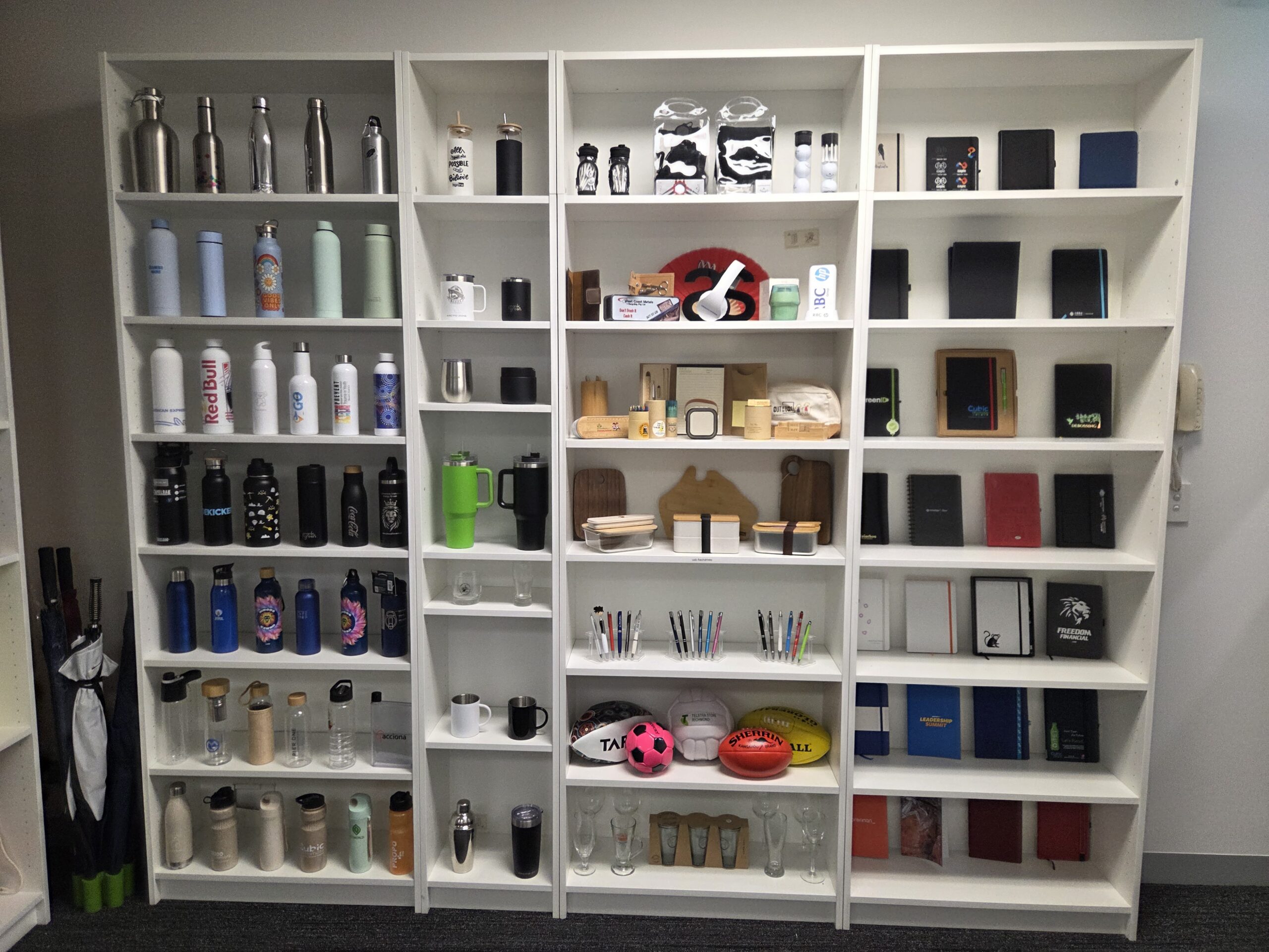 Three white shelves displaying merch samples—bottles, notebooks, mugs, and office items.