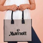 Person holding a beige tote bag with black Marriott logo and black handles.