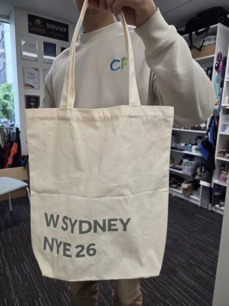Person holding a beige tote bag with W SYDNEY 2026—perfect for Aussie business needs.
