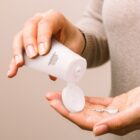 Person squeezing lotion from a white bottle onto their palm against a neutral background.