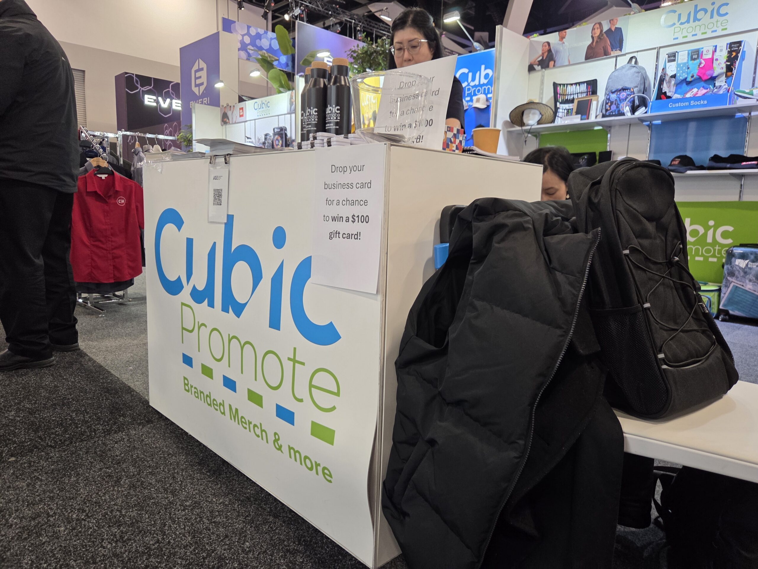 Booth with “Cubic Promote” branding, jackets on the counter, and products for trade shows displayed.