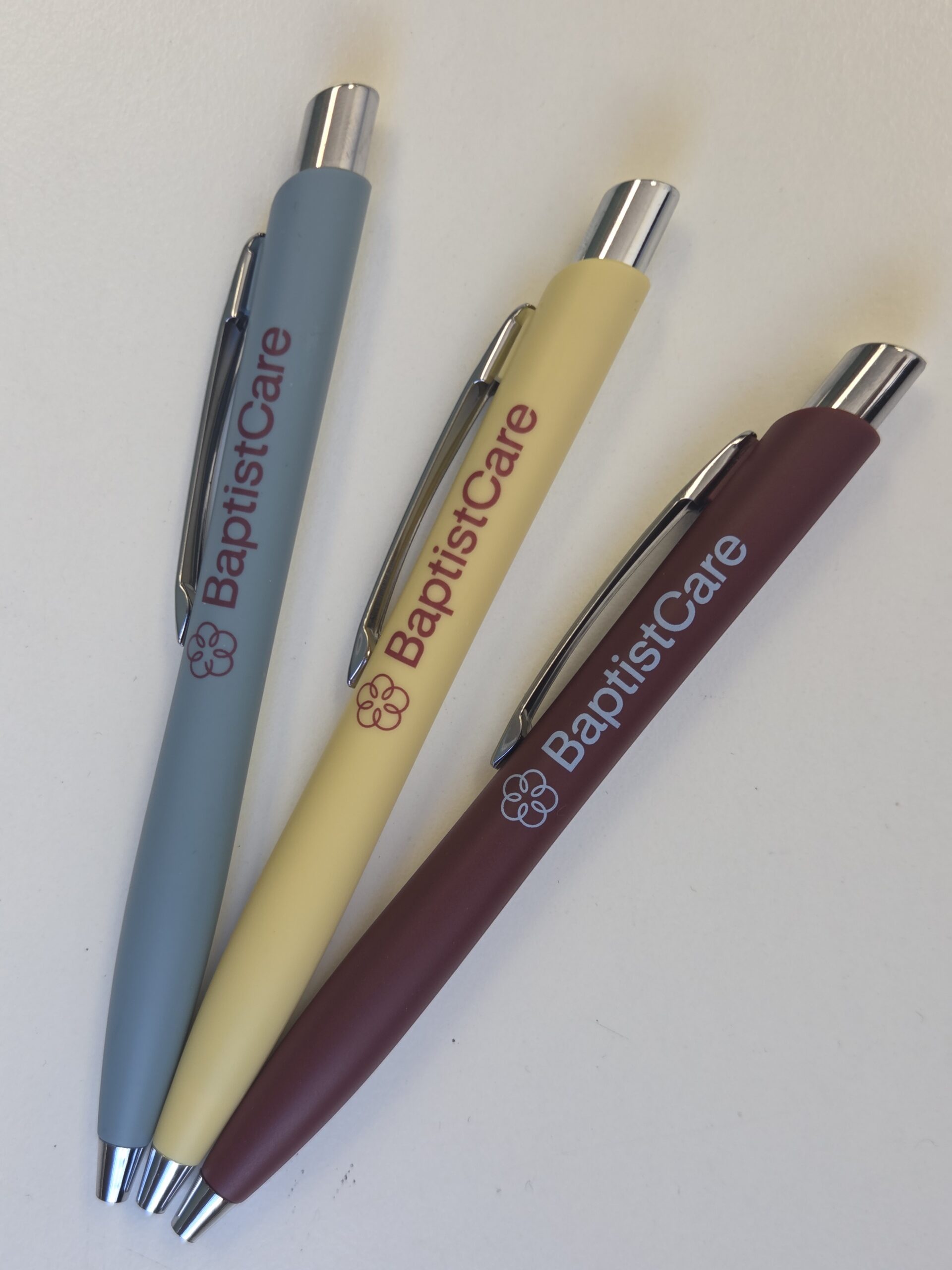 Three BaptistCare Event and Tradeshow Swag pens in blue, yellow, and maroon on a white surface.