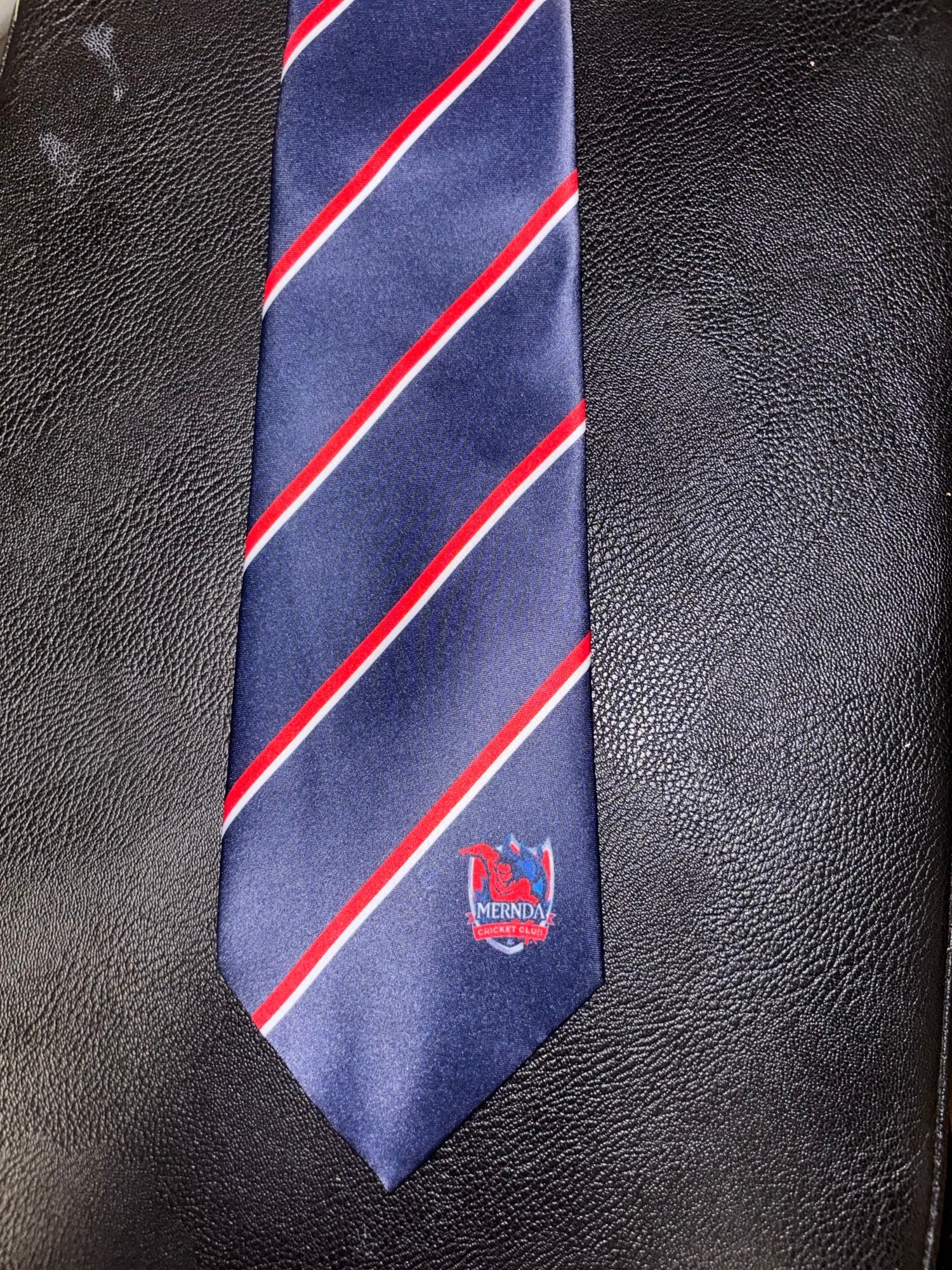 A navy blue tie with red and white stripes and a Mernda Cricket Club logo on a black surface.