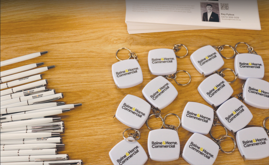 Pens, business cards, and Raine & Horne commercial real estate tape measure keychains on a table.