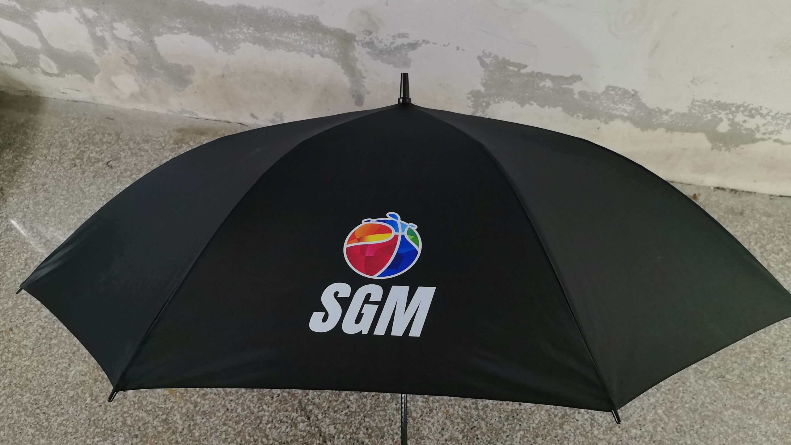 Black umbrella with a colorful globe logo and Sports Group Melbourne (SGM) text, indoors.
