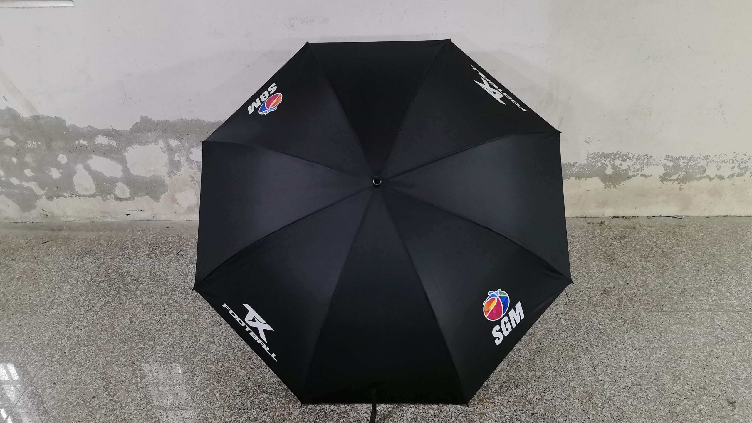 Black umbrella with colorful logos, Sports Group Melbourne text on gray speckled floor by a wall.