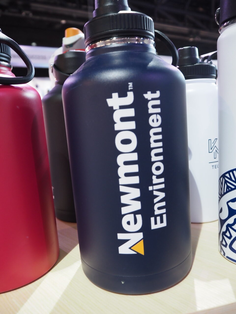 A black Newmont Environment bottle highlights bulk order benefits among other bottles on the table.