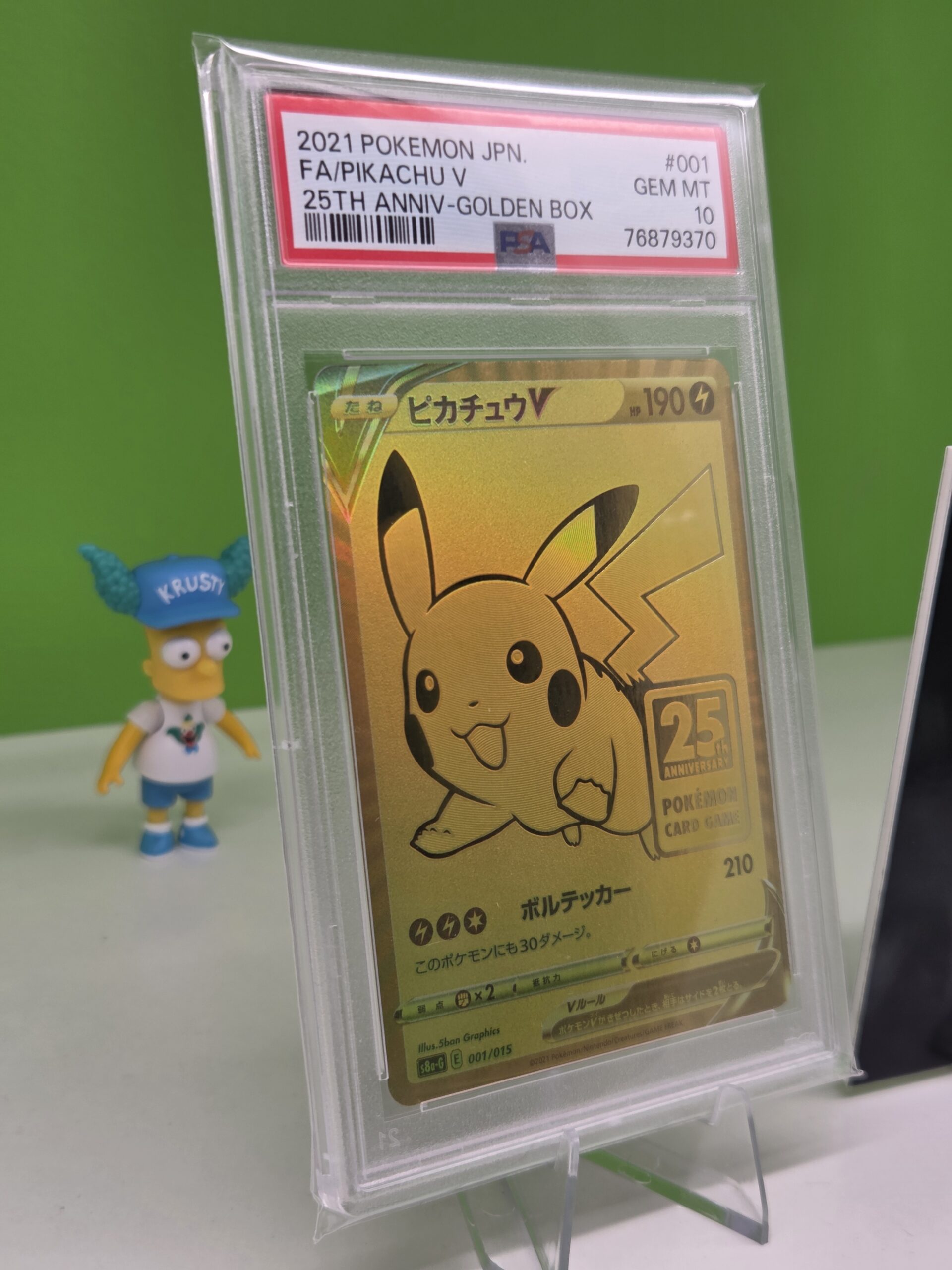 A graded gold Pikachu V Pokémon card in a pop-up display case, with a Bart figure nearby.
