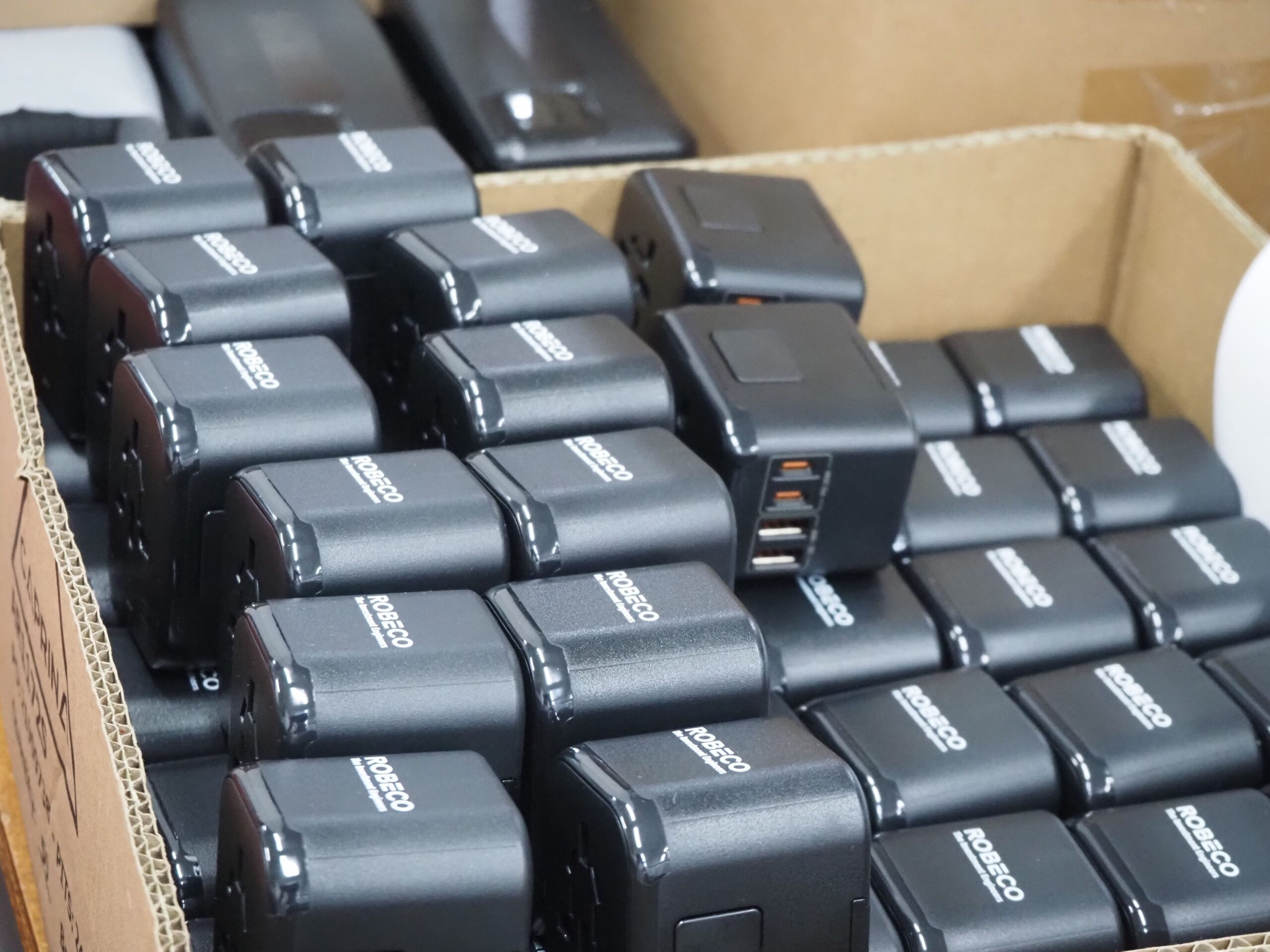 Black universal travel adapters, stacked for overseas production, fill a cardboard box.