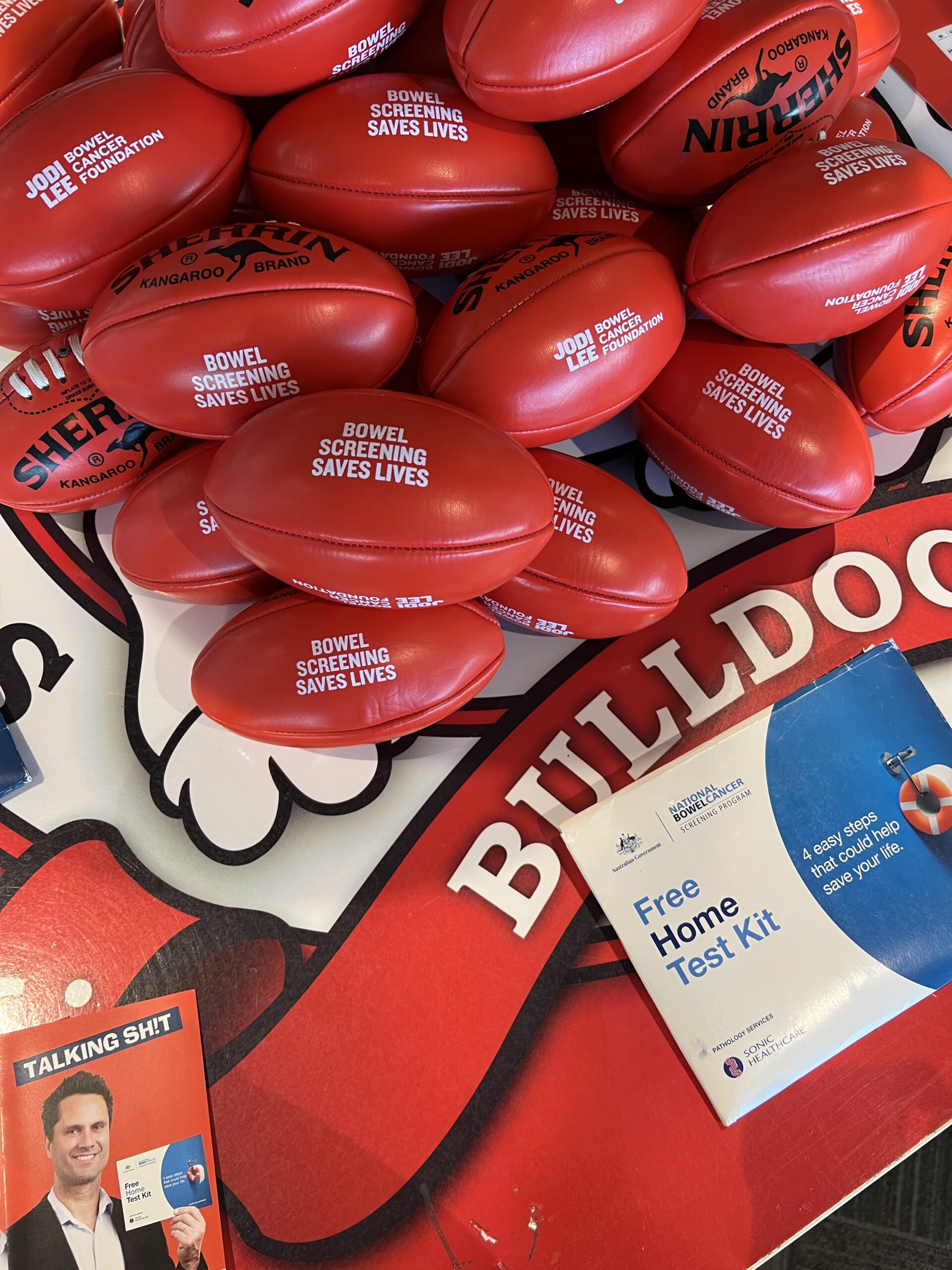 Red toy footballs with Bowel Cancer message and a Jodi Lee Foundation test kit on a table.