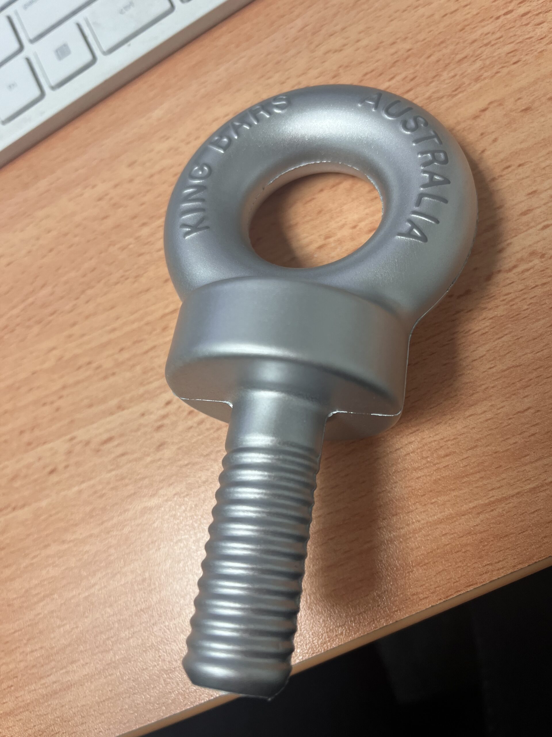 A large metal eye bolt labeled Australia from KingBars sits on a wooden desk near a keyboard.
