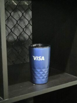 Blue travel mug with white VISA Australia logo on a dark shelf with a grid pattern behind.