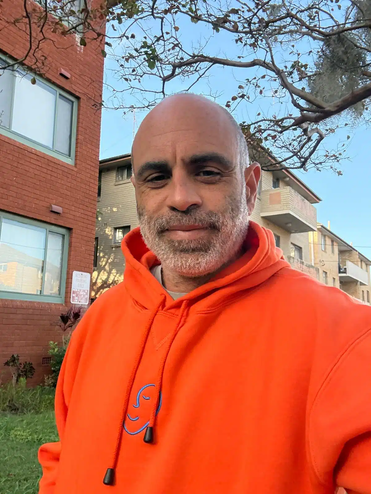 A man in an orange hoodie stands outside Sydney’s brick apartments and trees.