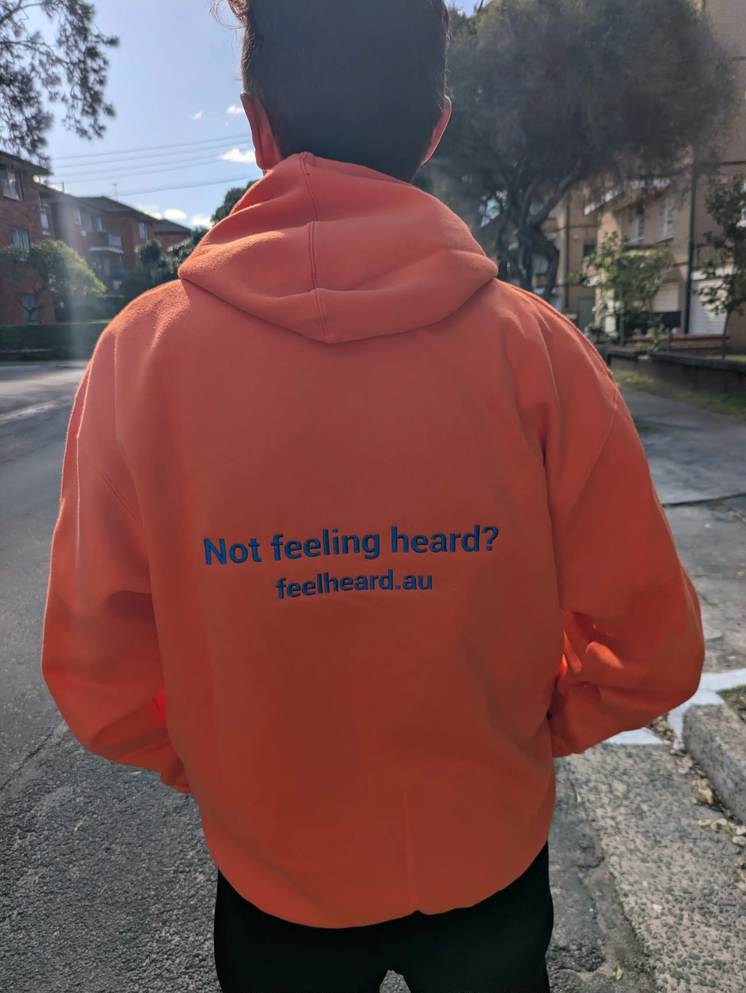 Person in orange hoodie with Feel Heard AU print on back, outdoors in Sydney.