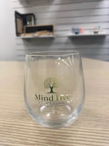 Clear stemless glass cup printed with MindTree Psychology full-colour tree logo, sitting on a timber desk.