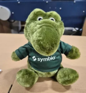 Custom green crocodile plush toy wearing a dark green Symbio branded t-shirt with white logo, made for Aussie Broadband.