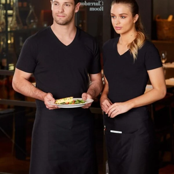 Two restaurant servers in black uniforms stand side by side. The man is holding a white plate with food, while the woman stands with her hands clasped. They are in a dining setting with tables in the background.