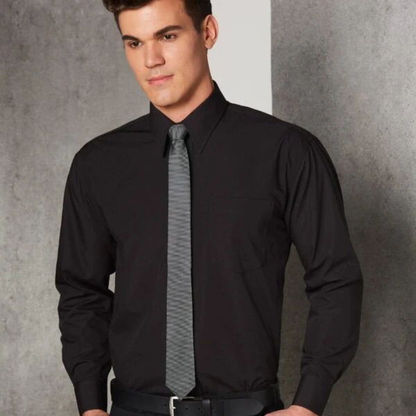 A person wearing a black dress shirt with a gray tie stands with hands in their pockets, leaning against a gray wall. The person has short dark hair and is looking to the side with a neutral expression.