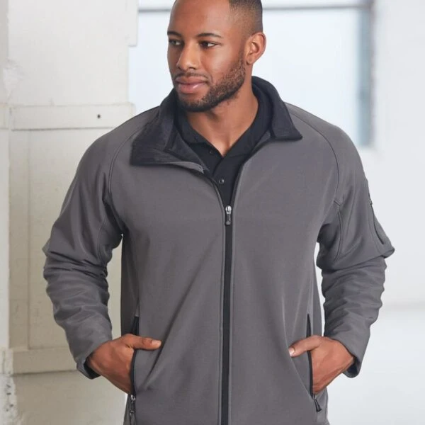 A man wearing a gray zip-up jacket with his hands in the pockets stands indoors. He has short hair and a trimmed beard, looking off to the side with a slight smile. The background features a white wall and a large window.