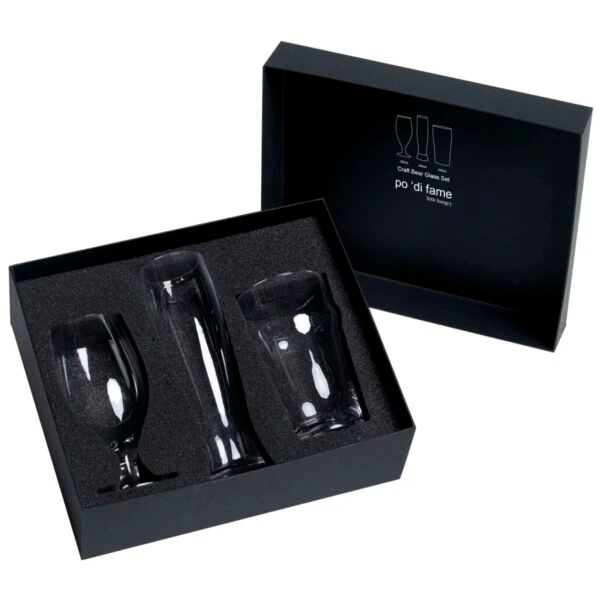 A black box contains three clear glass drinkware items fitted in a foam insert. The box lid, resting beside the glassware, has a design with outlines of the glasses and text indicating its a po di fame glass set.