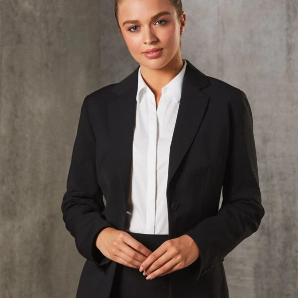 A woman with tied-back hair is wearing a black blazer over a white shirt. She is standing against a textured gray background, looking confidently at the camera with hands gently clasped.