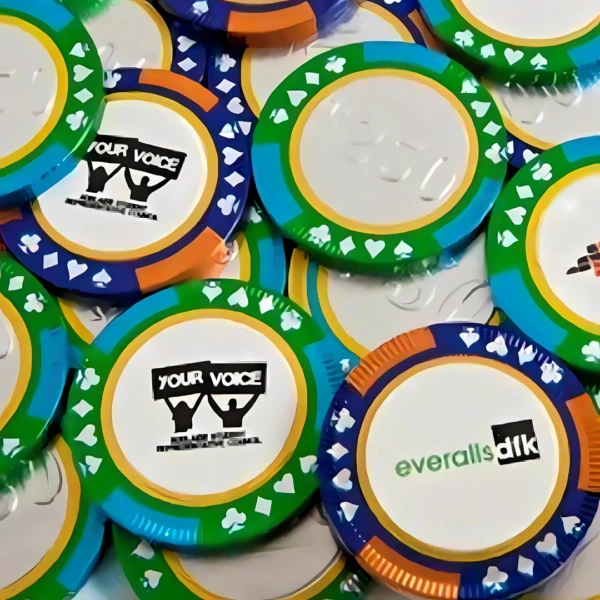 A close-up shows vibrant chocolate poker chips scattered on a surface. Some are personalized with YOUR VOICE, others display everall[s]ik. Resembling real poker chips, these colorful confections come in blue, green, and orange with card suit symbols around the edges.