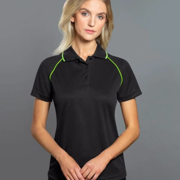 A woman with long hair wearing a black polo shirt with green piping stands against a gray background. She is looking directly at the camera with her hands clasped in front of her.