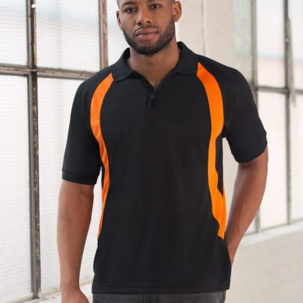 Person wearing a black polo shirt with orange stripes stands indoors near large windows, posing with one hand in pocket. Casual attire with a relaxed expression.