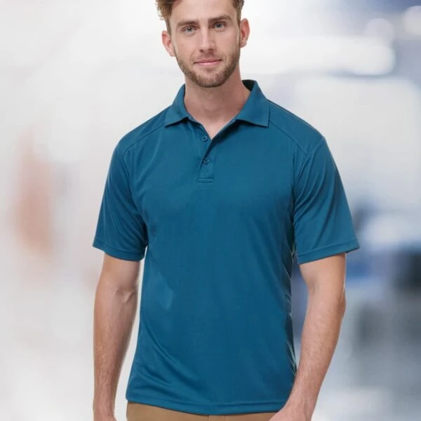 A man with short hair wears a teal polo shirt and tan pants. He stands against a blurred background, facing the camera with a neutral expression.