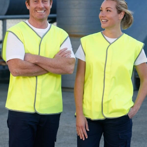 Two people wearing bright yellow safety vests and dark pants stand outdoors. The man has his arms crossed and is smiling, while the woman looks at him with a smile. They appear to be in a workplace setting with industrial equipment in the background.